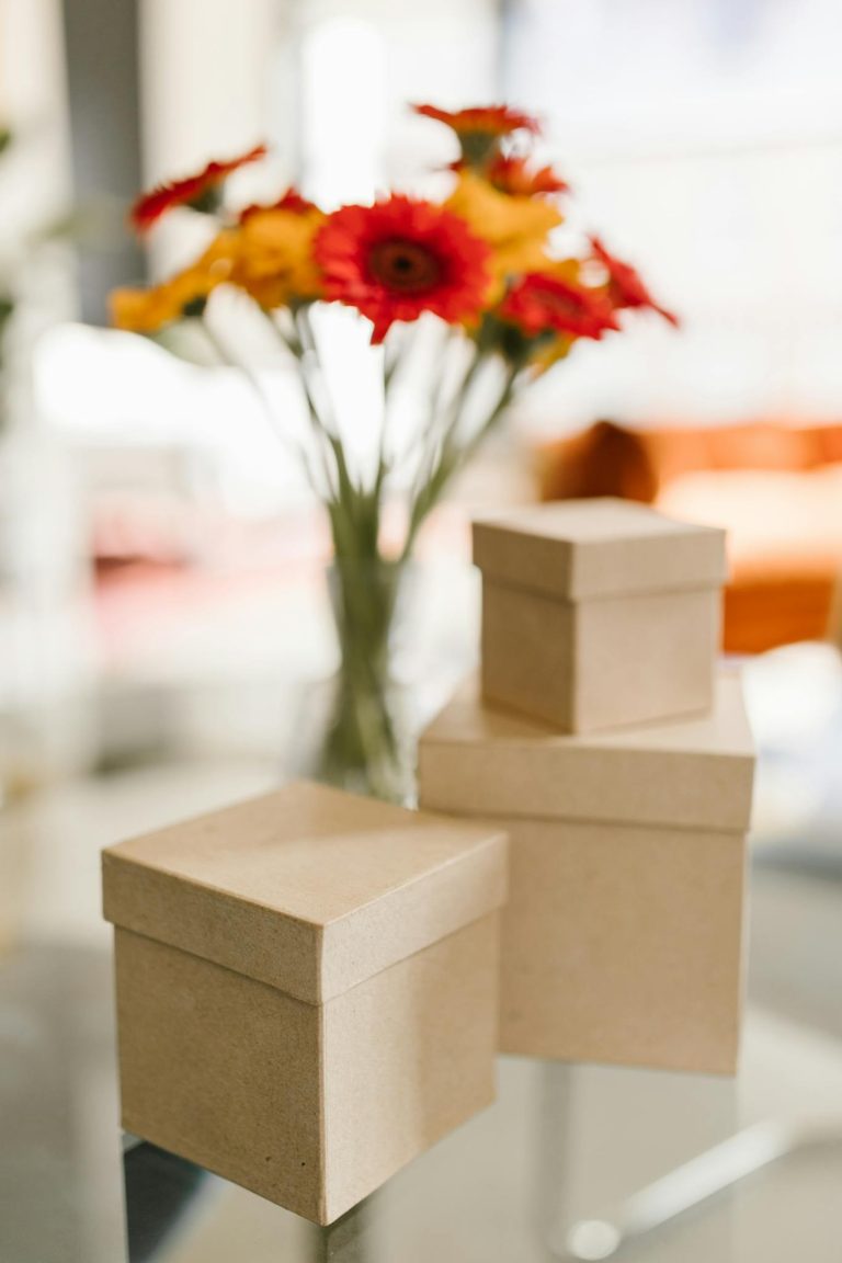 Minimalist shot of kraft boxes on a glass table with vibrant flowers in the background.